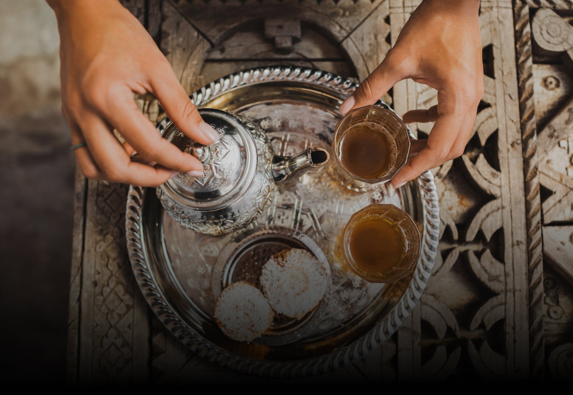 A person pours tea from a decorative teapot into a glass cup. The scene is set on an intricately carved wooden tray, which also holds another filled glass and three round pastries dusted with powdered sugar.