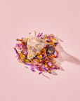 A Tea for Intuition: 1111: Pineapple & Butterfly Pea Flower Herbal Tea pyramid sachet resting on a loose pile of dried butterfly pea flower petals, yellow blossoms, and fruit pieces.