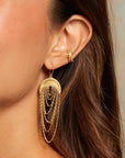 A close-up of a woman's ear showcases the Drape Chain Moon Earrings—18kt gold-plated earrings with layered chains hanging from a half-circle. Her brown hair is swept aside, revealing her smooth, glowing skin.
