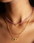 A woman in a white top wears three layered necklaces: a pink beaded strand, and two gold plated pieces—one being the Eye Blue Topaz Necklace with an eye-shaped pendant featuring blue topaz.