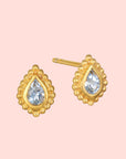 The Inner Light Blue Topaz Pear Stud Earring features a pear-shaped design with a clear blue topaz center, surrounded by a textured gold border and small round beads, inspired by delicate lotus motifs.