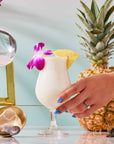A hand with blue nails holds a pina colada garnished with a pineapple slice and a purple flower—capturing the essence of summer refreshment. Nearby, a whole pineapple, a crystal ball, and a shimmering gold luxury tea infuser rest atop the glossy surface, evoking the elevated delights found in The Best of Summer Poolside Sips Box.