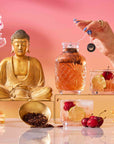 A hand holds a tea bag above a glass teapot beside a Buddha statue, flowers, bowls of Mai Tai: Black & Puerh Tropical Mocktail Tea, fruit cocktails, and cherries—all on a reflective surface with a pink-orange backdrop.