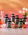 Seven jars of tea, featuring Summer Fridays™: Mai Tai, are displayed with a teapot, glassware, flowers, and fruit slices on a reflective surface. The scene evokes zero-proof cocktail elegance against a soft pink and orange gradient backdrop.