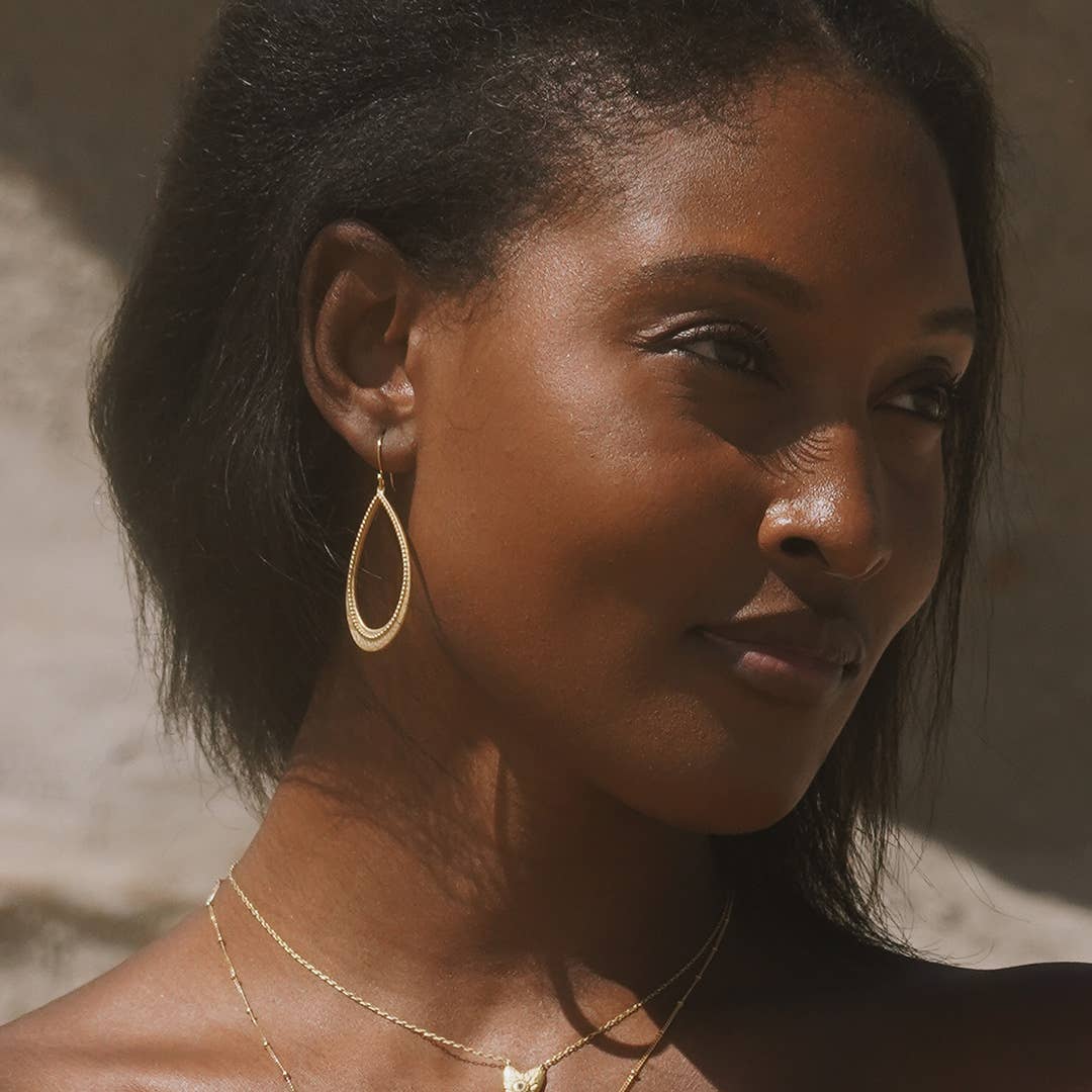 A woman with dark hair and smooth skin wears the Graceful Beauty Dot Teardrop Earrings in 18kt gold plate and layered necklaces, standing in soft sunlight against a neutral background.