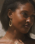 A woman with dark hair and smooth skin wears the Graceful Beauty Dot Teardrop Earrings in 18kt gold plate and layered necklaces, standing in soft sunlight against a neutral background.