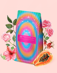 A vibrant tea package labeled "222: Hibiscus-Papaya Oolong with Roses" sits on a pink background, surrounded by roses, hibiscus, rosehips, and halved papaya to showcase its lively hibiscus and rose blend.
