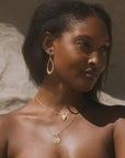 A woman with dark skin and straight black hair poses outdoors, wearing Graceful Beauty Dot Teardrop Earrings and two delicate gold necklaces—one with a pendant, the other with a coin charm. Sunlight highlights her face and shoulders.