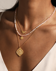 A woman in a white spaghetti strap top wears three layered necklaces: a pink and white beaded choker, a gold leaf charm, and an 18” White Topaz Lotus Medallion Necklace.