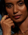A woman with medium skin tone and dark hair poses with her hand near her face, wearing the Emerald Radial Evil Eye Ring set of 3, along with gold earrings and a necklace, against a dark blue background.