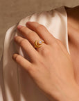 A hand rests on a shoulder, showcasing the Cradled in Light Moonstone Ring with a gold-plated crescent design and round white stone, set against a soft beige fabric background.