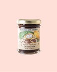 A jar from the Whipped Honey Gift Set by Savannah Bee Company is shown on a soft pink background. The label features cocoa beans and leaves, highlighting its delicious flavors with a net weight of 3 oz (85 g) noted at the bottom.