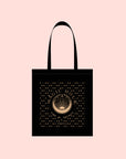 The Magic Hour Tote Bag, measuring 14x15 inches, features an intricate gold design of a central circle with rays and celestial symbols on a black cotton background. The light pink backdrop highlights the text "Magic Hour" with the smaller words "Time of Infinite," making it perfect for carrying during enchanted moments on the go.