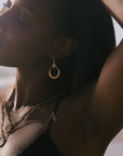 A woman with dark skin poses outdoors, eyes closed and sunlight highlighting her face, wearing a black top and the Stellar Sisterhood Moonstone Hoops with layered goddess jewelry.