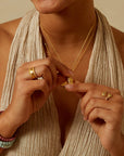 A woman wearing a beige textured halter top shows off the Inner Light I Am Loved Spinning Ring, layered gold necklaces, and colorful beaded bracelets, holding a small gold plated pendant between her fingers.