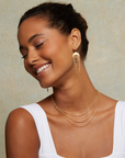 A woman with curly hair smiles with her eyes closed, wearing Drape chain moon earrings and layered gold necklaces, paired with a white sleeveless top against a neutral background.