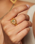 A hand showcases the Emerald Radial Evil Eye Ring set of 3, gold plated with green gemstones, worn on the ring finger. A white sleeveless top and a gold necklace are also visible in the background.