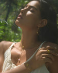A woman in a white dress stands outdoors with eyes closed, basking in sunlight. She wears gold earrings and the Transformed by Love Lotus Lariat Necklace. Green foliage forms a blurred background.