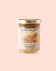 Against a pale pink background, a jar of Savannah Bee Company's 9 oz. Whipped Honey with Cinnamon, featuring a gold lid and label adorned with cinnamon sticks and leaves, is presented as part of their Whipped Honey Gift Set—an ideal addition for any tasting sample ensemble.