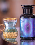 A close-up of a small, golden honeycomb-patterned glass cup with an elegant round handle sits next to a stylish dark bottle labeled "444: Sachiel's Prosperity," recognized for its organic ingredients. Both items rest on a reflective surface.