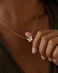 A woman wearing a ribbed olive-green top gently holds the Renewed Hope Pearl Butterfly Necklace, its 18k gold plated chain shimmering and highlighting the delicate mother of pearl butterfly pendant between her fingers.