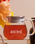 A glass Kinto One Touch Teapot filled with loose leaf tea, labeled "460ml," sits on a clear surface. The teapot has a metal lid with a built-in strainer. Behind the teapot, there is a blooming flower and an out-of-focus dark jar with an organic tea label on it.