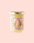 A 3-ounce jar from Savannah Bee Company's Whipped Honey Gift Set features a beige label with a beehive and flowers illustration, gold lid, and is elegantly displayed against a pale pink background.