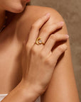 A woman gently rests her hand on her shoulder, highlighting the Cradled in Light Moonstone Ring—gold plated with a crescent moon design and round white stone—set against a neutral brown background.