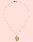 The Gold Buddha Pendant Necklace features a delicate 18kt gold-plated chain with a round, intricately engraved Buddha coin pendant, shown on a white background.