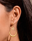 A close-up of a woman’s ear wearing the Stellar Sisterhood Moonstone Hoops—gold crescent moon earrings with a small sun design above the crescent. Her straight, dark hair is tucked behind her ear.