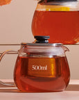 A clear glass Kinto Stainless Steel Teapot from Magic Hour, with a black lid, holding 500ml of amber-colored loose leaf tea, sits on a reflective surface. Another similar teapot is partially visible in the background, positioned on an elevated surface. Both teapots feature metal infusers.