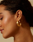 A woman with curly dark hair in a low bun wears several gold earrings, including a large textured hoop, small studs, and Inner Light Blue Topaz Pear Stud Earrings—symbolizing new beginnings—set against a neutral background. Her skin glows softly.