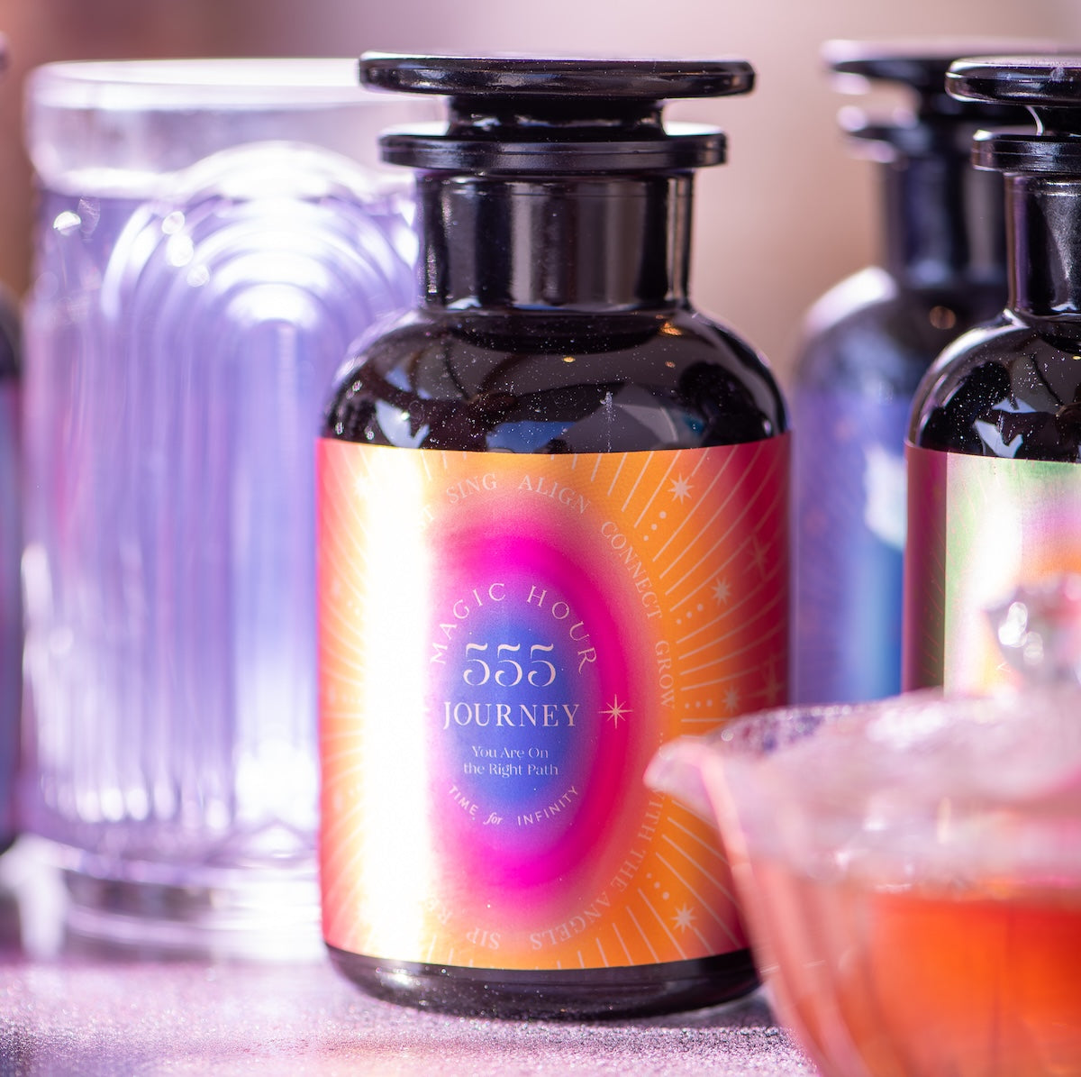 A close-up of a dark glass bottle with a vibrant "555" label, reading "Journey" and "You Are On the Right Path," is surrounded by softly blurred bottles. The background has a warm glow and a partially visible glass dish.