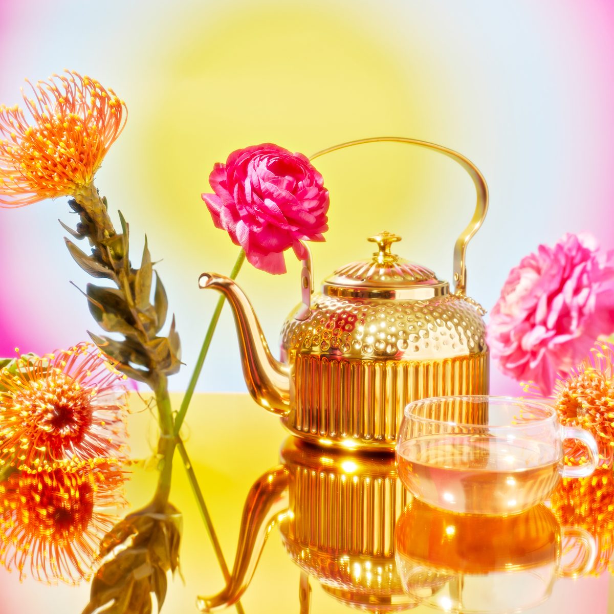 The Celestial Harmony set includes a golden teapot and transparent teacup on a reflective golden surface, surrounded by vibrant pink and orange flowers. A soft yellow to blue gradient in the background creates a warm atmosphere that evokes the serene aura of spiritual awakening.