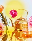 The Celestial Harmony set includes a golden teapot and transparent teacup on a reflective golden surface, surrounded by vibrant pink and orange flowers. A soft yellow to blue gradient in the background creates a warm atmosphere that evokes the serene aura of spiritual awakening.