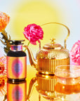 A vibrant arrangement showcases the 555 golden teapot alongside a glass teacup brimming with tea on a reflective surface. Nearby, a jar with the colorful 555 label is encircled by vivid pink and orange flowers, set against a bright gradient backdrop.