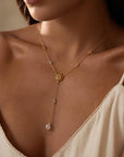 A woman in a cream spaghetti strap top showcases the Transformed by Love Lotus Lariat Necklace, an 18k gold plated Y-necklace with a sun-shaped pendant and small gemstones, featuring a teardrop stone at the end.
