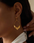 Close-up of a woman wearing Fluttering Petals Gold Earrings, large and modern with a curved leaf design in 18kt gold plating. She touches her neck and wears a white top with visible back button details.