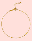 The 9" Shimmering chain anklet + 1" extender chain features delicate 18kt gold plating, tiny textured beads spaced along a shimmering chain, an adjustable clasp, and a small circular charm at the end.