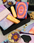 A flat lay of the Beauty Inside & Out Box featuring a botanical serum, cosmic-themed cards, dried flowers, and a black massage tool, all artfully arranged on a purple and peach backdrop.