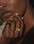 A person with dark skin gently touches their face, showcasing two gold rings—one with a dark, oval stone and the Cradled in Light Moonstone Ring—against a softly lit background.