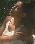 A woman in a cream dress sits outdoors, eyes closed in sunlight. She wears gold jewelry, including the Transformed by Love Lotus Lariat Necklace, with matching bracelet, ring, and earrings, gently resting her hand on her shoulder amid greenery.