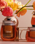 Glass teapots filled with amber-colored Magic Hour Kinto Stainless Steel Teapot are displayed. The largest teapot, labeled "720ml," sits on the left with another teapot to the right on a metal stand. Bright flowers in a vase are in the background, adding a colorful contrast.