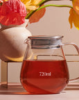 A Kinto One Touch Teapot marked "720ml" filled with red liquid, possibly Magic Hour Tea, sits on a table. Behind it, there is a ceramic vase holding vibrant pink and yellow flowers. The scene is well-lit with a soft, warm light.