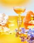 A glass of orange liquid is surrounded by a white rose, an orange poppy, and blue flowers on a reflective surface. This echoes the charm of our Flower Power Mother's Day Mystery Box, set against a warm yellow gradient radiating bliss.