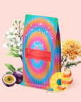 A vibrant, rainbow-colored coffee bag labeled "777: Celestial Messengers Dawn Pineapple Passionfruit Green Tea" sits among a chrysanthemum, flowering branch, sliced passionfruit, and spiral-cut orange on a pastel pink background.