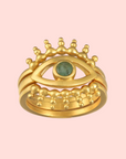 The Emerald Radial Evil Eye Ring set of 3 features gold-plated eye-shaped rings with green gemstone irises and gold bead accents resembling eyelashes, displayed against a pale pink background.