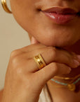 A close-up of a woman's face and hand shows her wearing the Inner Light I Am Loved Spinning Ring, paired with gold plated hoops and layered necklaces. Her lips are slightly parted as she smiles naturally, resting her chin on her hand.