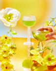 A hand holds an 888: Golden Gateway cocktail—green with golden citrus—amid bright yellow flowers and a white blossom. In the background, another 888: Golden Gateway sits on a tall, white stand under sunny abundance frequency.