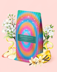 A vibrant tea bag showcases a rainbow mandala, white flowers, apple slices, bananas, and vanilla beans on soft pink. The label reads "888: Lemon-Jasmine-Vanilla Herbal Tea," inspired by Archangel Michael’s prosperity blend.